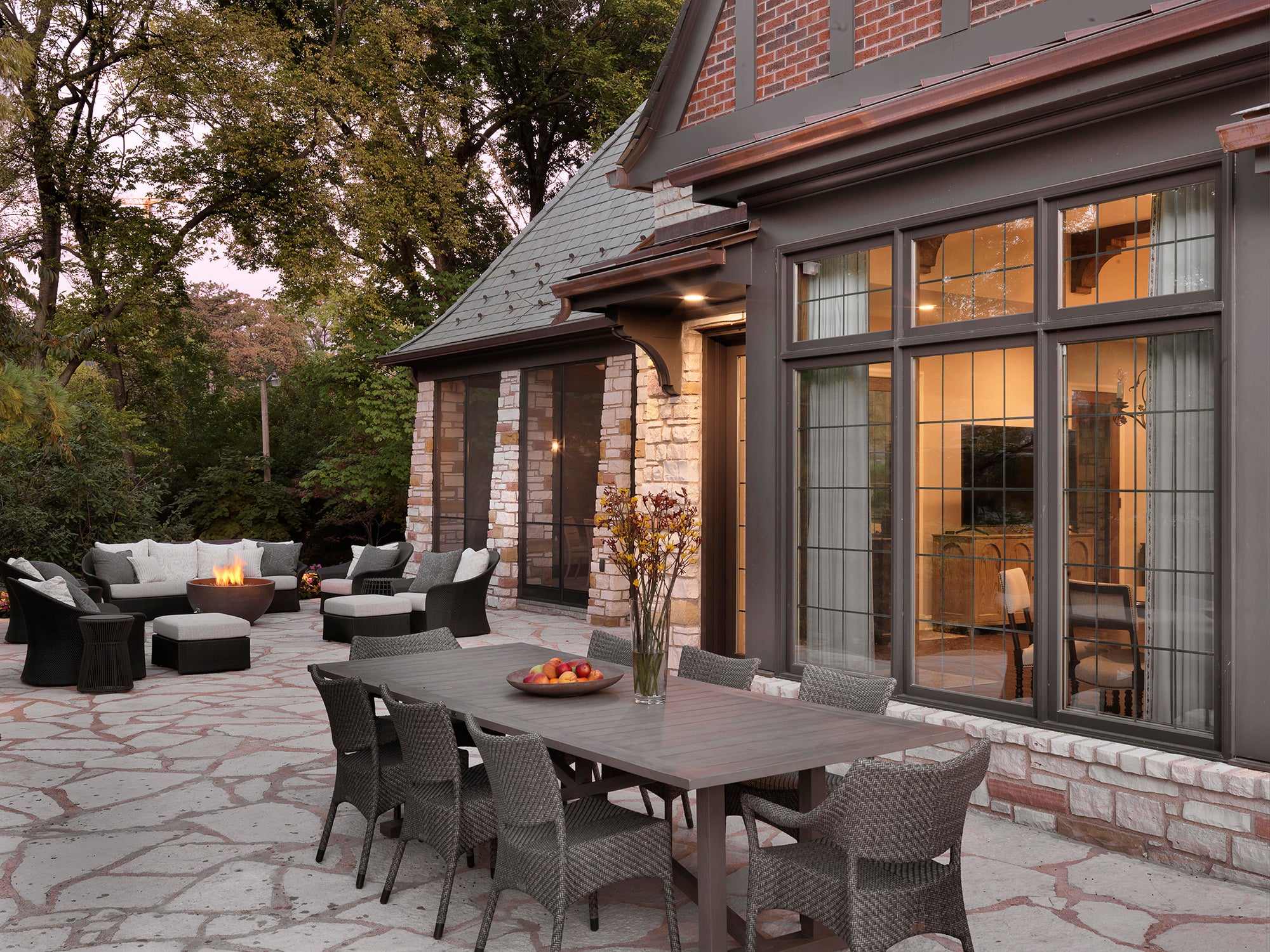 Outdoor patio at 1920s Clayton Estate with long stone table, white seating, dark accents, and fire feature at dusk — luxury design by Ättlingar.